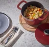 A cast iron pot filled with food is placed on a wooden board on a speckled countertop, next to a plate and cutlery. Produktbild-Vorschau