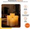 Four LED candles of varying heights are arranged on a wooden surface, with a blurred background of glassware and foliage. Produktbild-Vorschau