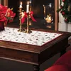 A Christmas-themed table runner with a festive pattern is laid out on a dark wooden table, adorned with two brass candlesticks holding lit candles and a vase of red flowers. A fireplace and a decorated Christmas tree are visible in the background. Náhľad obrázku produktu