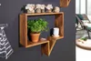 A wooden wall shelf with a plant and jars on it, set against a dark gray wall. A lounge chair and a round rug are visible in the background. Produktbild-Vorschau