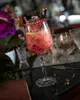 A cocktail in a stemmed glass on a dark reflective surface, with flowers and a book in the background. Produktbild-Vorschau