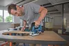A man is using a cordless jigsaw to cut a curved shape into a piece of wood on a workbench. Anteprima dell’immagine prodotto