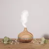 A wooden essential oil diffuser emitting mist, placed on a wooden surface with greenery and small white flowers around it. Podgląd zdjęcia produktu