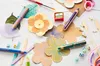 A collection of art supplies, including pencils, a sharpener, and paper flowers, arranged on a white surface. Voir les images du produit