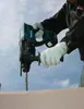 A person wearing white gloves is using a teal and black rotary hammer drill to drill into a light brown surface, with a cloudy blue sky in the background. Produktbild-Vorschau