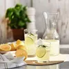 Two glasses of lemonade with lime and rosemary, a carafe of lemonade, and a plate of lemons on a counter. Produktbild-Vorschau