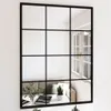 A mirror with a black frame is mounted on a white wall. The mirror reflects a living room scene with a sofa, pillows, and a table with books and plants. Podgląd zdjęcia produktu