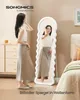 A woman is standing in front of a full-length mirror in a bedroom setting. Produktbild-Vorschau