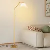 A gold floor lamp with a pleated white shade is positioned next to a cream-colored sofa with decorative pillows. Podgląd zdjęcia produktu