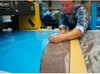 A man in a workshop is cutting a piece of material next to a large blue sheet. Produktbild-Vorschau