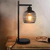 A black metal table lamp with a spiral cage shade sits on a wooden table. Podgląd zdjęcia produktu