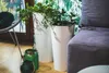 Two white planters with green plants are placed on a wooden floor next to a gray sofa and a chair. There are also two decorative green lanterns in the foreground. Anteprima dell’immagine prodotto