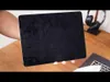 A person is holding a black laptop or tablet case over a wooden desk. Headphones are visible in the background. Produktbild-Vorschau