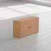 A cork yoga block is placed on a light-colored wooden floor, with sunlight casting shadows from a window. Produktbild-Vorschau