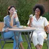Two women are sitting at a table outdoors, each holding a portable blender and drinking from it. There is a bowl of chips and dip on the table. Podgląd zdjęcia produktu