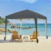 A dark gray pop-up canopy tent is set up on a sandy beach with two wooden chairs and a small table in front of it. The ocean and rocky coastline are visible in the background. Produktbild-Vorschau