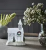 A decorative Christmas church figurine is displayed on a table, with flowers in a vase in the background. Produktbild-Vorschau