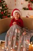 A young child wearing a Santa hat and a red sweater is sitting on a couch, wrapped in a festive blanket with a snowman pattern. A decorated Christmas tree is visible in the background. Náhled obrázku produktu