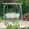 A hammock swing chair with cushions is hanging from a wooden pergola in a garden setting. There is a pile of firewood to the right and plants in the foreground. Voir les images du produit