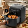 A person's hand is holding the handle of a black air fryer filled with cooked chicken. The air fryer is on a wooden table with other food items in the background. Produktbild-Vorschau
