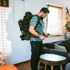 A man is putting on a large backpack in a living room. Produktbild-Vorschau
