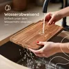 A person is rinsing a wooden cutting board under a faucet in a kitchen sink. Produktbild-Vorschau