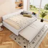 A white daybed with a pull-out trundle bed is set up in a room with a large window overlooking a green landscape. Decorative pillows and a patterned comforter are on the daybed. A rug with a geometric pattern is on the wooden floor. Náhled obrázku produktu