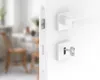 A close-up view of a white door handle and lock mechanism, with a blurred background of a dining area. Produktbild-Vorschau