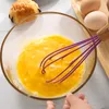 A hand is whisking eggs in a glass bowl with a purple whisk. There are two eggs and a pastry in the background. Produktbild-Vorschau
