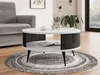 A modern living room scene featuring a round coffee table with a slatted base, placed on a circular rug. A sofa is visible to the left, and a window with greenery is in the background. Produktbild-Vorschau