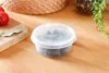 A round, transparent plastic container filled with small, dark items sits on a wooden surface. To the left, a cutting board holds two red tomatoes and a bay leaf. In the background, a woven mat and a plant are visible. Náhled obrázku produktu
