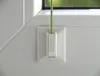 A close-up view of a white plastic window accessory with a green cord running through it, attached to a white window frame. Produktbild-Vorschau