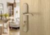 A close-up of a bronze door handle with keys inserted, set against a wooden door. The background shows a blurred interior of a room. Produktbild-Vorschau