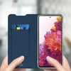 A person is holding a blue flip cover phone case with a credit card inserted, and a smartphone with a colorful floral wallpaper is visible. Náhled obrázku produktu