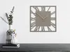 A modern square wall clock with Roman numerals and wooden hands is displayed on a white wall above a dark wooden surface. To the left, a decorative vase with eucalyptus branches and a smaller white vase with cotton flowers are placed on the surface. Náhled obrázku produktu