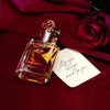 A bottle of perfume is placed on a red velvet cloth next to a rose and a handwritten note. Podgląd zdjęcia produktu