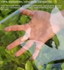 A hand is shown through a transparent material, with green foliage in the background. Voir les images du produit