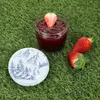A jar of strawberry jam with a strawberry on top, two whole strawberries, and a decorative lid, all placed on a grassy surface. Anteprima dell’immagine prodotto