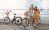 Two women are standing next to electric bikes on a boardwalk, eating ice cream. Produktbild-Vorschau