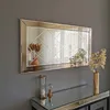 A rectangular mirror with a decorative frame hangs on a gray wall above a mirrored console table. The table holds an hourglass and a vase with dried flowers. Another vase with dried flowers is reflected in the mirror. Produktbild-Vorschau
