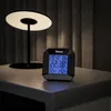 A digital clock sits on a nightstand next to a modern lamp. Anteprima dell’immagine prodotto