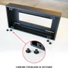 A black metal housing for a table fireplace is shown on a white surface, with a close-up inset of rubber feet. Náhľad obrázku produktu