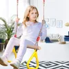 A young girl is swinging indoors on a wooden trapeze swing with yellow plastic rings. Produktbild-Vorschau