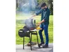 A man is grilling food on a charcoal smoker grill in a backyard setting. Produktbild-Vorschau