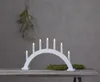 A white arched candle holder with seven lit bulbs sits on a wooden table, with a sprig of greenery hanging from the ceiling behind it. Produktbild-Vorschau