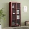A tall, bordeaux red display cabinet with glass doors stands against a light green wall, next to a large potted plant. Decorative items are visible inside the cabinet. Náhľad obrázku produktu
