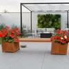 An outdoor patio scene with a wooden bench featuring integrated planters filled with red flowers, a pergola, and seating area in the background. Náhled obrázku produktu