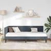 A dark gray velvet corner bed frame with a white mattress and pillows, set against a white wall with two wooden shelves displaying decorative items and books. A floor lamp is visible on the left, and a large potted plant on the right. Produktbild-Vorschau