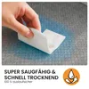 A hand is holding a white pad over a textured gray and blue surface, demonstrating absorbency. Produktbild-Vorschau
