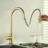 A gold kitchen faucet with a pull-out spray head is shown in use, with water running into a sink containing vegetables. Produktbild-Vorschau